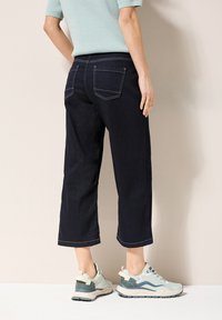 Mörk denim, mid-rise capribyxor med synliga bakfickor och kontrasterande sömmar, ihop med ljusa sneakers med texturerade detaljer.