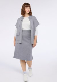 Grauer Strickcardigan mit kurzen Ärmeln, kombiniert mit einem taillierten grauen Rock und weißen Sneakers. Das Material wirkt weich und glatt.