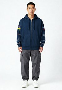 Sweat à capuche zippé marine avec poches avant. Présente des broderies colorées sur les manches, des manches élastiques et une coupe décontractée, associé à un pantalon cargo gris.