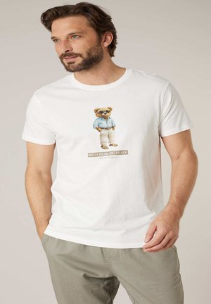 Homme portant un t-shirt blanc avec un graphique d'un ours en lunettes de soleil, chemise bleue, pantalon beige et le texte "Rich Bear, Big Flair", debout les mains dans les poches.
