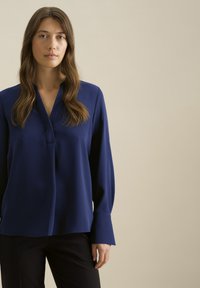 Blusa blu navy con colletto rialzato, maniche lunghe e patta frontale. Realizzata in tessuto liscio, presenta una vestibilità morbida e dettagli minimi.