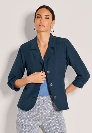 Vrouw draagt donkerblauw blazer over lichtblauw topje en wit-blauwe broek met patroon, poseert met hand die blazer rechtzet.