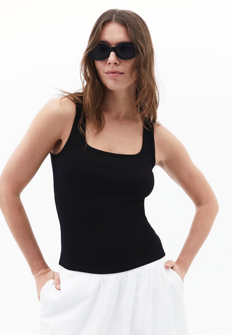 Débardeur noir à côtes avec un décolleté carré. Le mannequin porte des lunettes de soleil noires ovales et un pantalon blanc ample avec des poches.