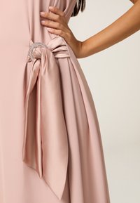 Abito rosa chiaro dalla forma fluida, con un accento a nodo in raso fissato da un dettaglio in strass in vita. Texture liscia ed elegante.