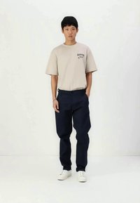 Bež majica s kratkimi rokavi s tiskanim logotipom, združena s temno mornarsko širokimi hlačami in belimi supergami.