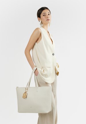 Donna con blazer crema senza maniche e pantaloni che tiene una grande borsa tote beige con portachiavi decorativo, in piedi davanti a uno sfondo bianco.