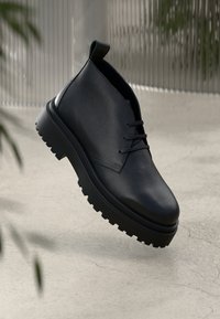 Μαύρη δερμάτινη μπότες στον αστράγαλο με υφασμάτινη επιφάνεια, σχέδιο με κορδόνια και χοντρή ελαστική σόλα με έντονη υφή για πρόσφυση.