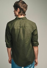 Chemise olive à boutons avec une texture douce, manches retroussées et ourlet arrondi. Le détail du pli au dos ajoute une touche de design subtile.