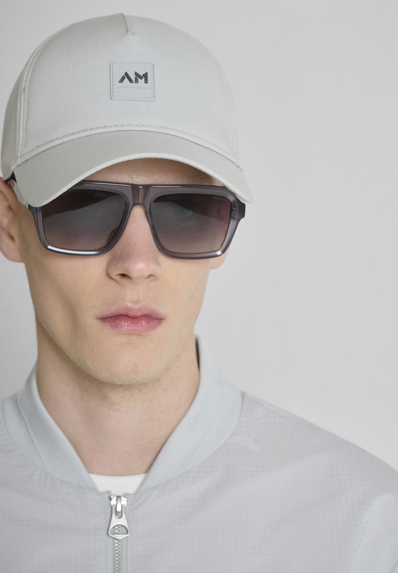 Jonge man met een lichtgrijze pet met logo, grote donkere zonnebril en een lichtgrijze jack met rits voor een effen achtergrond.