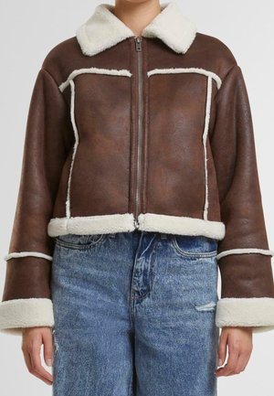 Brązowa kurtka ze skóry owczej z białą podszewką z polaru, z zamkiem błyskawicznym z przodu, szerokimi mankietami, noszona z niebieskimi jeansami z wysokim stanem i małą rozdarciem na lewym udzie.