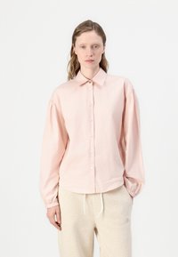 Light pink button-up shirt with long sleeves, gathered shoulders, and a collar. Made from a soft fabric with a subtle sheen and minimal logo detail.