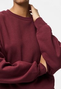 Pessoa com um sweatshirt de manga longa bordô, com uma mão a tocar o pescoço e o outro braço cruzado sobre o corpo.