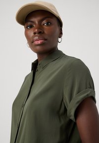 Olivenfarget skjorte med knapper og rullede ermer, kombinert med en lys tan-cap. Stoffet ser glatt ut med et beskjedent kragedesign.