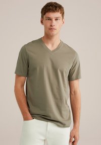 Olivno zelena v-neck majica iz bombažnega materiala, kratki rokavi, sproščen kroj, nosi se z belimi hlačami. Gladka tekstura, sproščen dizajn.