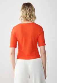 Haut à manches courtes en tissu texturé orange vif. Présente une encolure large et se ajuste près du corps, associé à un pantalon de couleur claire.