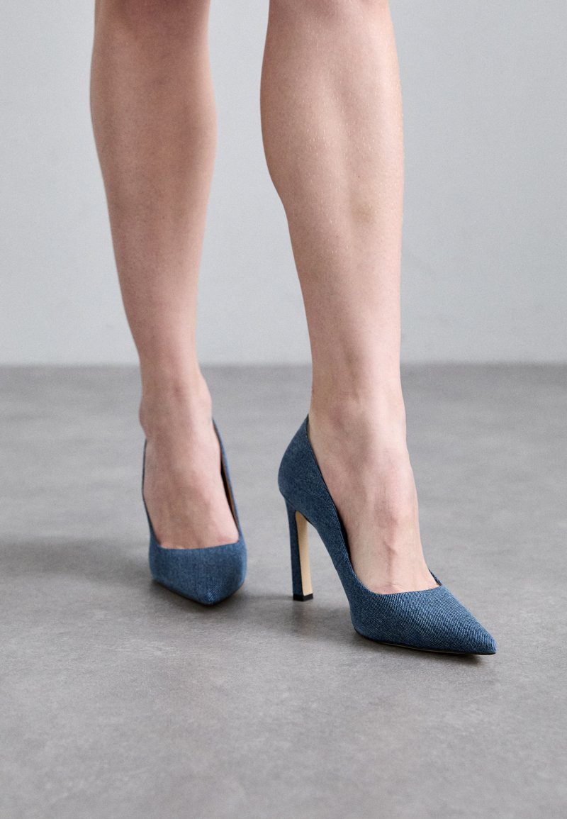 Blå denim højhælede pumps med spids tå og åbne sider, der har en stiletto hæl og en glat, tekstureret stoffinish.