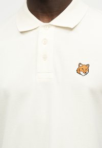 Polo blanco con tres botones y un logo bordado de la cabeza de un zorro naranja en el pecho izquierdo, llevado por una persona de piel oscura.