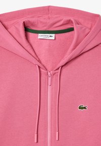 Haut à capuche zippé rose avec doublure intérieure verte. Présente une texture de tissu douce et un petit logo crocodile vert sur la poitrine. Coupe classique.