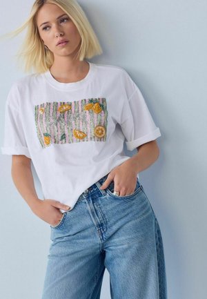 Femme blonde portant un T-shirt blanc avec une broderie de fruits jaunes et un jean bleu, debout les mains dans les poches contre un mur clair.