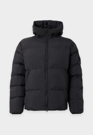 Un modèle pose devant un fond clair uni, vêtu d’une doudoune noire à capuche, d’un pantalon de jogging crème avec le logo EA7 et de baskets blanches.