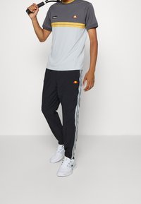 Osoba v šedém a žlutém sportovním tričku, černých teplákových kalhotách se bočními pruhy, bílých teniskách a držící tenisovou raketu.