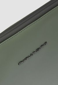 Groene pouch van synthetisch materiaal met een gladde textuur en een zwarte rits. Aan één zijde een verhoogd, zwart logo.