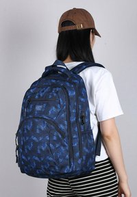 Zaino blu navy con un motivo in tessuto testurizzato, dotato di più compartimenti con zip e spallacci regolabili. Abbinato a una camicia bianca e pantaloni a righe.