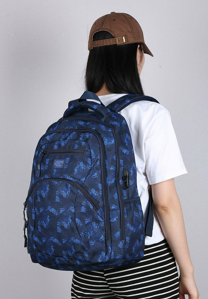 Zaino blu navy con un motivo in tessuto testurizzato, dotato di più compartimenti con zip e spallacci regolabili. Abbinato a una camicia bianca e pantaloni a righe.