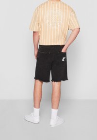 Mustat farkkushortsit, joissa on riipitettu helma ja takataskut, yhdistettynä vaaleankeltaiseen, raidalliseen lyhythihaiseen paitaan, jossa on pyöreä graafinen kuvio.