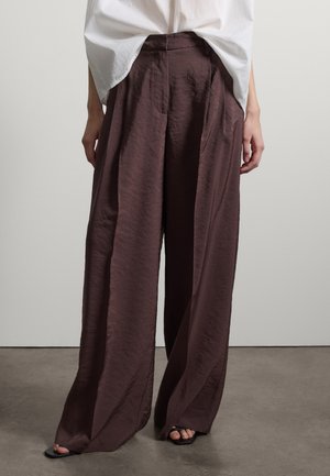 Person trägt lose, weit geschnittene dunkelbraune Hose mit weißem Oberteil, das hineingesteckt ist, und steht auf einem grauen Boden vor einer einfachen weißen Wand.