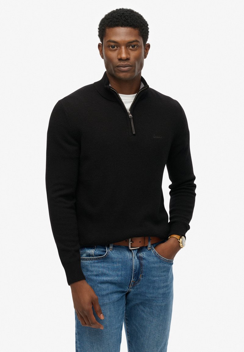 Jersey negro con cremallera en el cuello, de manga larga, con puños acanalados y un acabado texturizado, llevado sobre una camisa blanca, con jeans azules y un cinturón marrón.