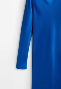 Robe bleue vif à manches longues suspendue sur un fond blanc, montrant le haut du bras et une partie du torse.