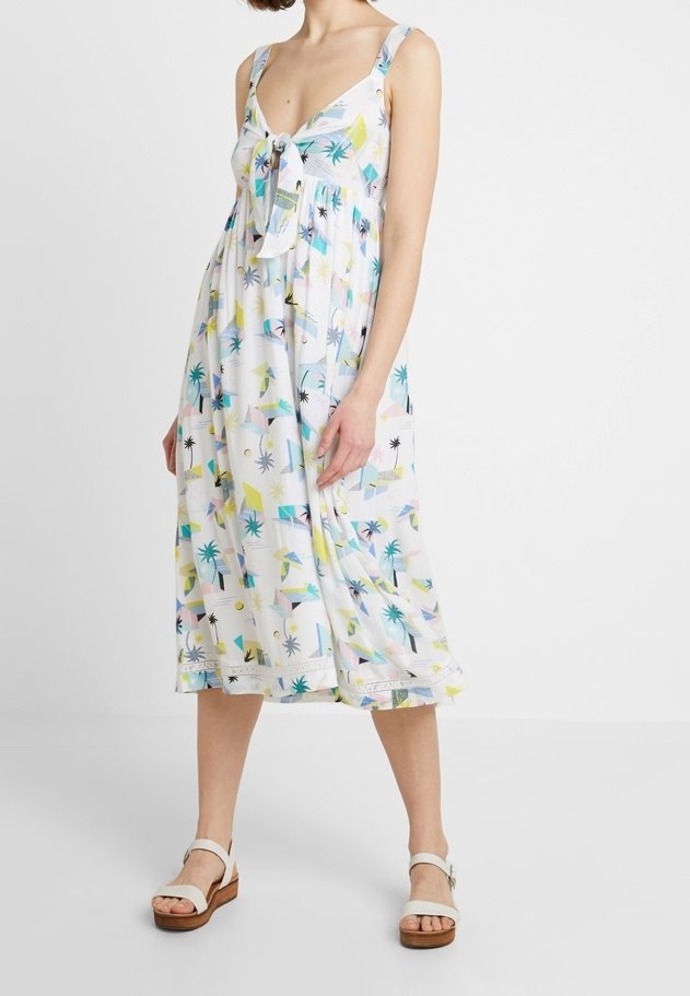 Femme portant une robe midi blanche sans manches avec des motifs géométriques colorés et des palmiers, et des sandales blanches à plateforme.