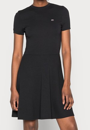 Frau trägt ein schwarzes Kleid mit kurzen Ärmeln und einem kleinen rechteckigen Logo auf der linken oberen Brust, steht vor einem einfarbigen Hintergrund.