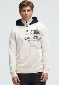 Weißer Strickpullover mit hohem Kragen und marineblauer Kapuze, ausgestattet mit einer Knopfleiste und aufgedruckten Logos auf der Vorderseite. Strukturierter Stoff sichtbar.