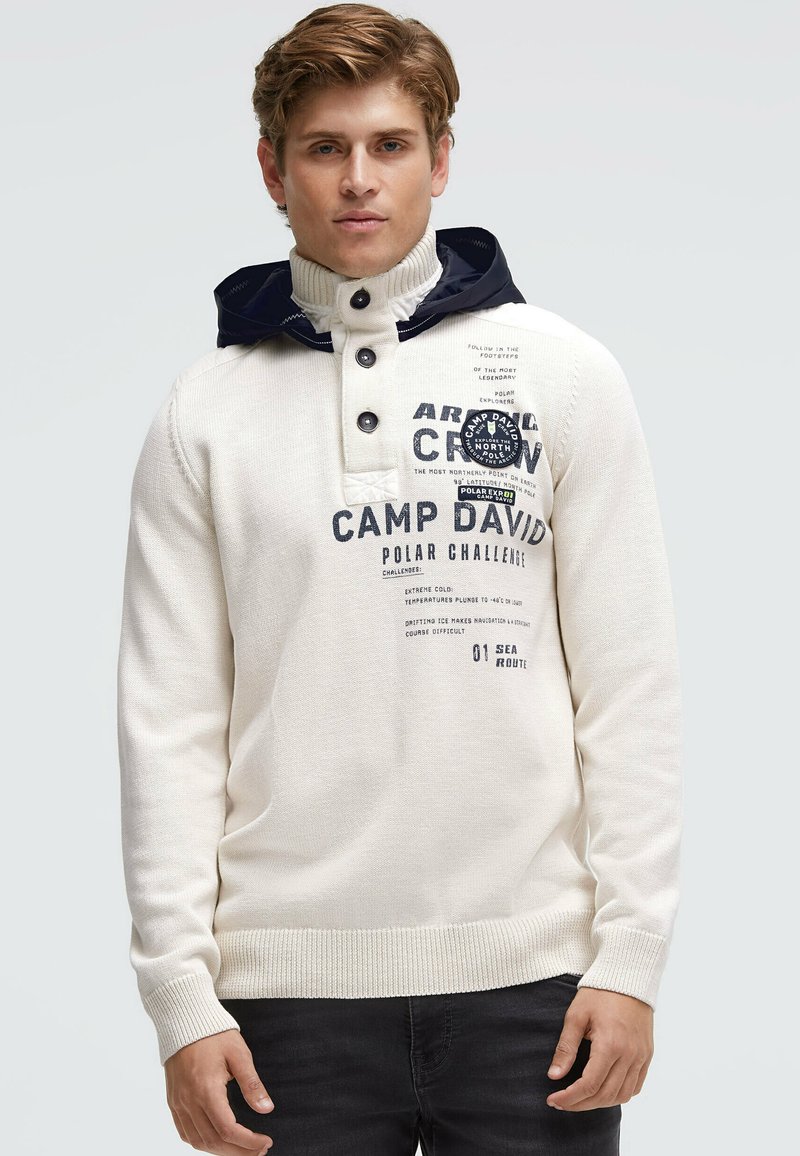 Weißer Strickpullover mit hohem Kragen und marineblauer Kapuze, ausgestattet mit einer Knopfleiste und aufgedruckten Logos auf der Vorderseite. Strukturierter Stoff sichtbar.