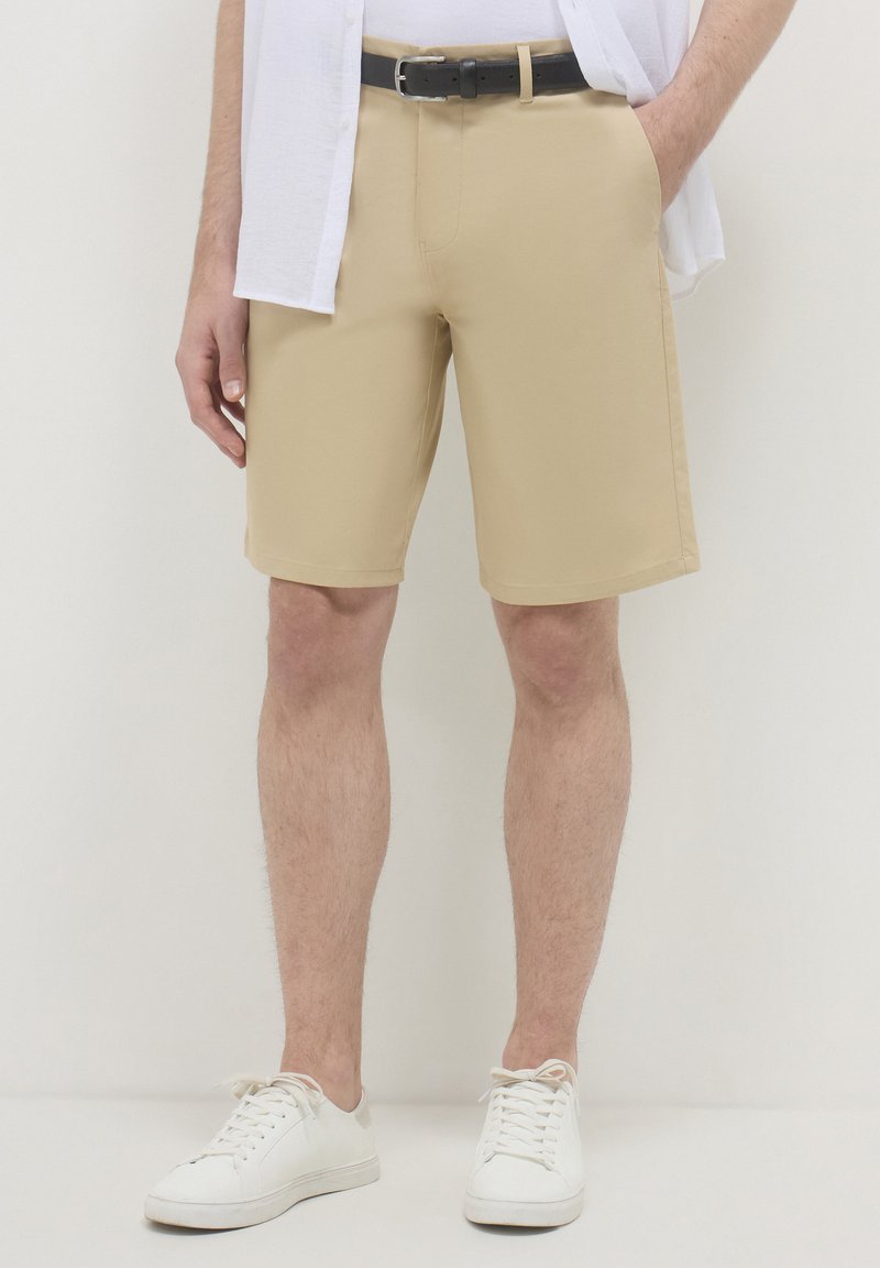Mann trägt beige, knielange Shorts mit einem schwarzen Gürtel, weiße Sneaker und ein teilweise sichtbares weißes Hemd.