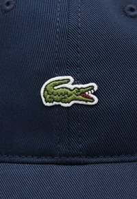 Cappellino blu navy realizzato in tessuto testurizzato, con un logo di coccodrillo ricamato in verde e accenti rossi sul lato.