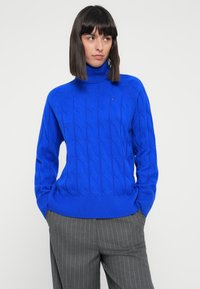 Blue cable-knit turtleneck sweater with ribbed hem and cuffs, featuring a small logo on the chest, worn with gray pinstripe pants.