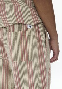 Compleu din bumbac cu dungi verticale în nuanțe de bej, roșu și marron. Detalii notabile includ o buzunar pe pantaloni și un etichetă de mărime vizibilă.