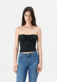 Top corto negro sin mangas con detalle de nudo en la parte delantera, hecho de una tela suave y elástica. Combinado con jeans azul de cintura alta.