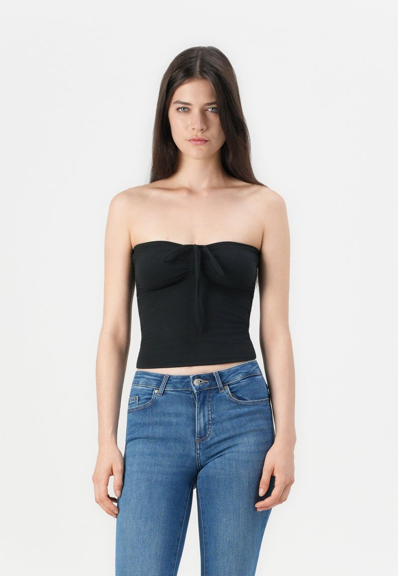 Top corto negro sin mangas con detalle de nudo en la parte delantera, hecho de una tela suave y elástica. Combinado con jeans azul de cintura alta.