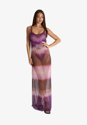 Donna che indossa un vestito lungo, senza maniche, semi-trasparente con sfumatura viola e rosa, in piedi con una mano sul fianco.