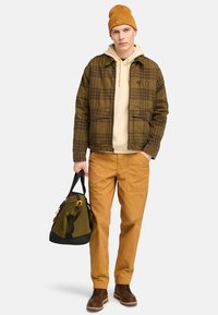 Brunternet skjortejakke, beige hætteklædte trøje, tan bukser og brune støvler. Holder en mørkegrøn og sort duffel taske med orange detaljer.