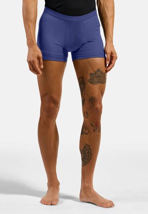 Male model wearing blue boxer briefs, standing barefoot with multiple black tattoos on right leg against a plain light background.