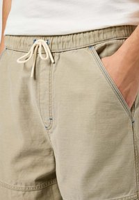 Beżowe bawełniane szorty z elastycznym pasem i sznurkiem. Wyróżniają się niebieskimi przeszyciami na kieszeniach oraz gładką fakturą.