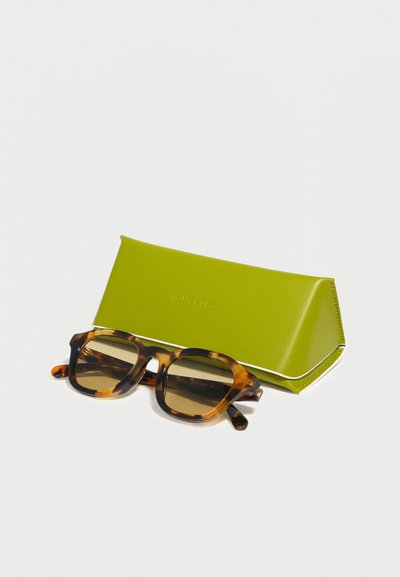 Lunettes de soleil à monture écaille avec des verres bruns placées à côté d'un étui rigide triangulaire vert sur une surface blanche.