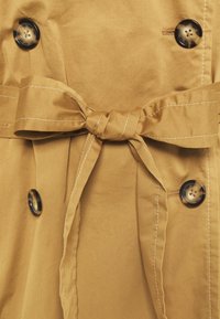 Beżowy płaszcz trench z materiału z przewiązanym paskiem, z ciemnymi guzikami i widocznymi detalami ściegów. Gładka struktura i dopasowany fason.