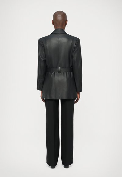 Čierny kožený blejzer s dlhými rukávmi a opaskom v páse, skombinovaný s vysokými čiernymi nohavicami. Hladká textúra a prispôsobený strih.