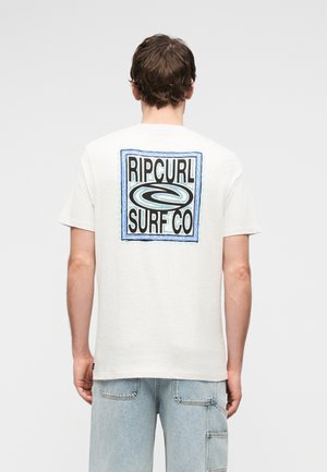 Ein Mann von hinten, der ein weißes T-Shirt mit einem blau umrandeten Rip Curl Surf Co Logo auf dem Rücken und hellblaue Jeansshorts trägt.